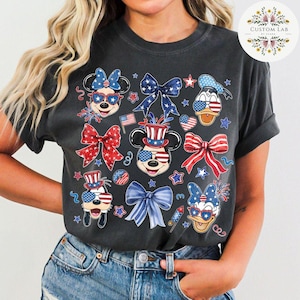 Peut inclure: Un t-shirt gris fonc&eacute; sur le th&egrave;me patriotique avec Mickey Mouse, Minnie Mouse, Donald Duck et Dingo portant des chapeaux et des lunettes de soleil rouge, blanc et bleu. Le t-shirt est d&eacute;cor&eacute; d'&eacute;toiles, de n&oelig;uds et de feux d'artifice.