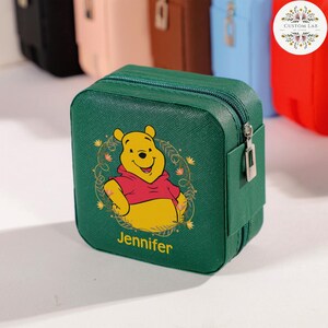 May include: A green, square jewelry case with a zipper closure. The case features a cartoon image of Winnie the Pooh in a red shirt, with the name "Jennifer" printed below. Other colored cases are visible in the background.
