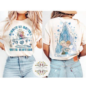 Puede incluir: Camiseta color crema con un dise&ntilde;o tem&aacute;tico de Disney. La parte delantera muestra a Elsa y Anna en una taza de t&eacute; con el texto "Disney is Better With Sisters". La parte trasera muestra a las hermanas abraz&aacute;ndose frente a un castillo con el texto "Custom Name."
