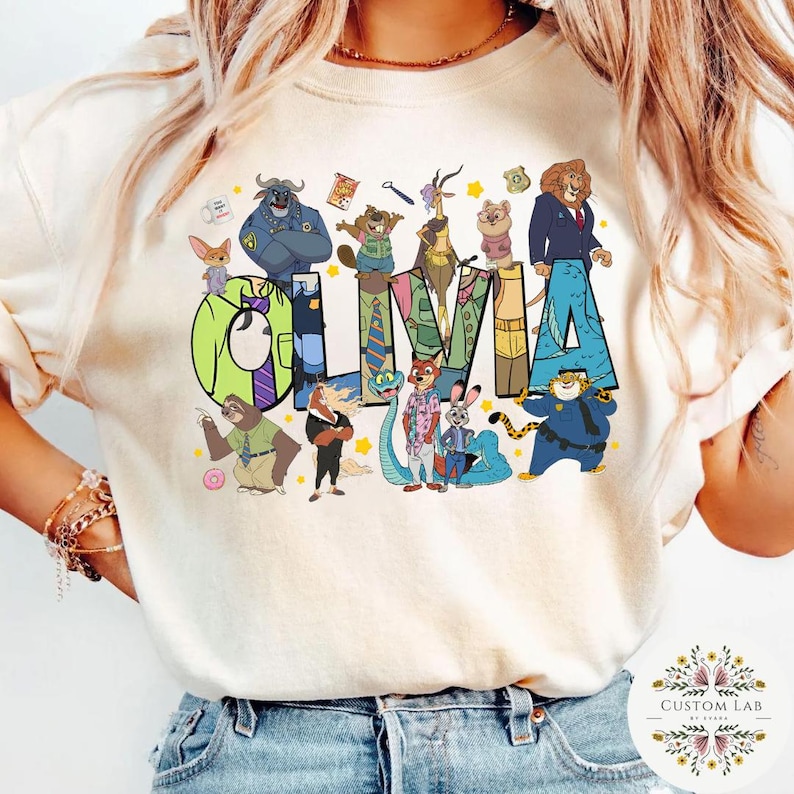 Pu&ograve; includere: Felpa color crema con il nome "Olivia" a grandi lettere colorate, circondata da personaggi dei cartoni animati di un popolare film d'animazione. Il design include vari personaggi di animali ed elementi decorativi.