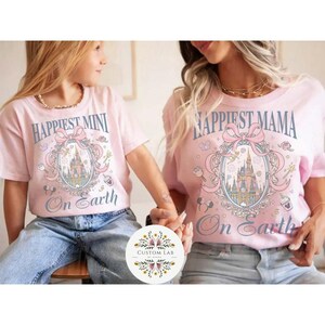 May include: Two light pink t-shirts with matching designs. One says "Happiest Mini On Earth" and the other says "Happiest Mama On Earth". Both shirts feature a castle graphic with a pink bow and decorative elements.