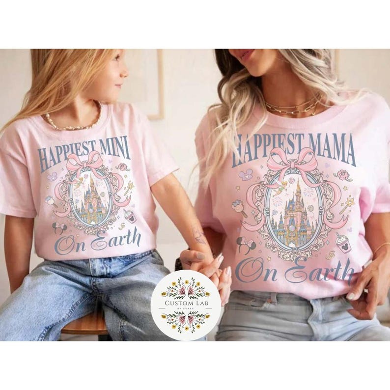 May include: Two light pink t-shirts with matching designs. One says "Happiest Mini On Earth" and the other says "Happiest Mama On Earth". Both shirts feature a castle graphic with a pink bow and decorative elements.