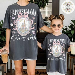 May include: Two matching charcoal gray t-shirts. One says "Happiest Mama On Earth" and the other says "Happiest Mini On Earth". Both shirts feature a castle design with a pink bow and decorative elements.