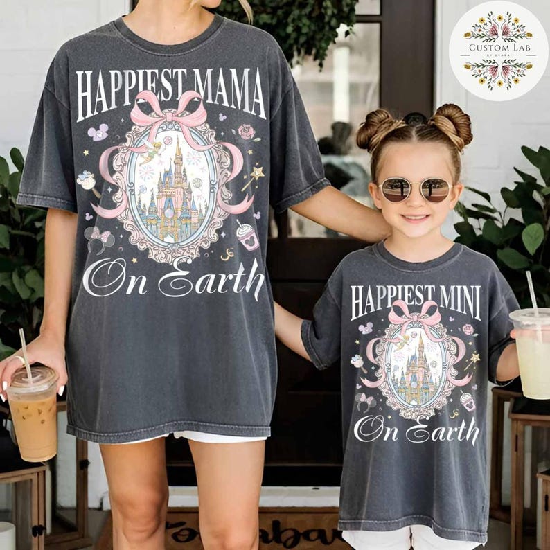 May include: Two matching charcoal gray t-shirts. One says "Happiest Mama On Earth" and the other says "Happiest Mini On Earth". Both shirts feature a castle design with a pink bow and decorative elements.