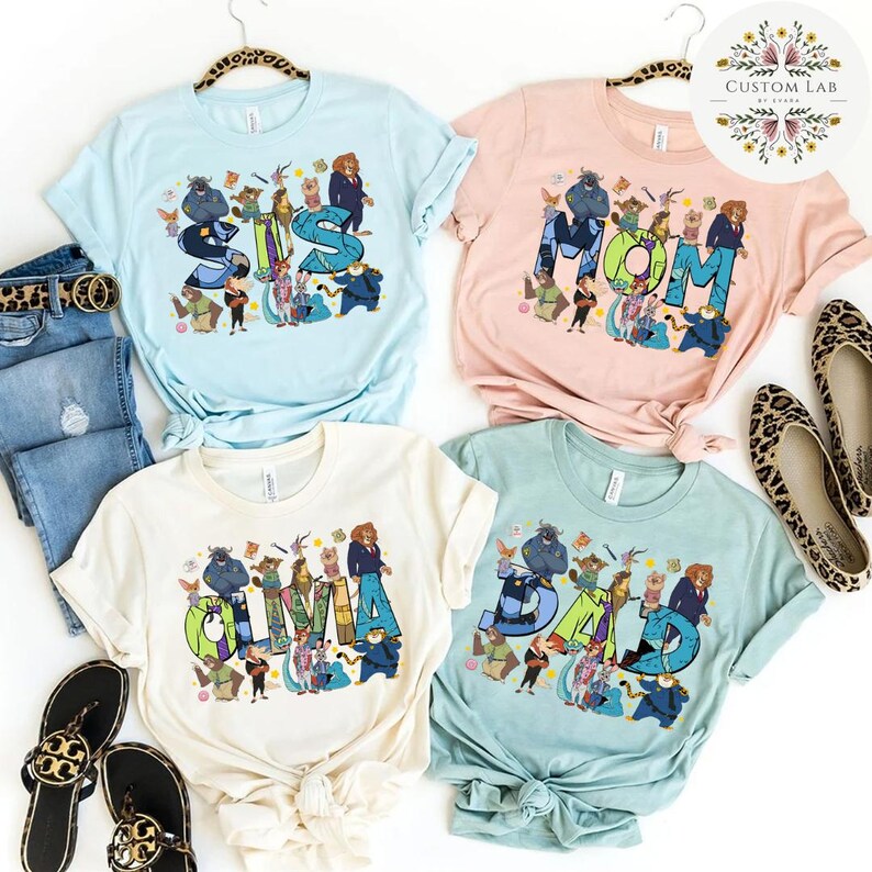 Pu&ograve; includere: Quattro t-shirt grafiche in azzurro, rosa, crema e verde acqua. Ogni maglietta presenta una parola diversa: "SIS", "MOM", "OLIVIA" e "DAD", con illustrazioni di personaggi dei cartoni animati. Le magliette sono esposte con jeans, scarpe e accessori.