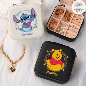 May include: Square, zippered jewelry cases featuring Disney characters Stitch and Winnie the Pooh. The Stitch case is off-white with a floral wreath and the name "Jennifer". The Winnie the Pooh case is black with a floral wreath and the name "Jennifer". A gold heart pendant necklace is also visible.