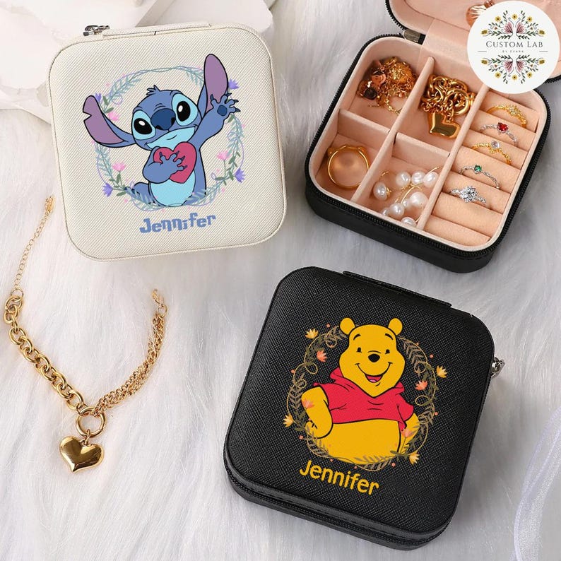 May include: Square, zippered jewelry cases featuring Disney characters Stitch and Winnie the Pooh. The Stitch case is off-white with a floral wreath and the name "Jennifer". The Winnie the Pooh case is black with a floral wreath and the name "Jennifer". A gold heart pendant necklace is also visible.