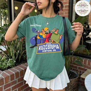 May include: A green t-shirt with a graphic featuring characters from the movie "The Emperor's New Groove." The shirt has the text "runDisney" and "Kuzcotopia Virtual 5K" with the year 2026. The shirt is paired with a white pleated skirt.