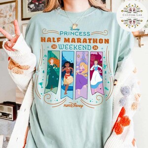 May include: A light blue t-shirt with the text "Disney Princess Half Marathon Weekend 20-26". The shirt features illustrations of Disney princesses on vertical banners. The "runDisney" logo is at the bottom.