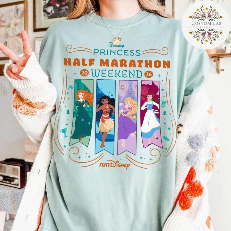 May include: A light blue t-shirt with the text "Disney Princess Half Marathon Weekend 20-26". The shirt features illustrations of Disney princesses on vertical banners. The "runDisney" logo is at the bottom.