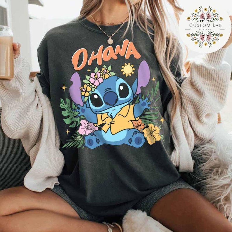 May include: Dark gray t-shirt featuring a cartoon character with the word "OHANA" above it. The character is blue with large ears, wearing a flower crown and yellow shirt. Tropical flowers and leaves surround the character. The shirt is a casual style.
