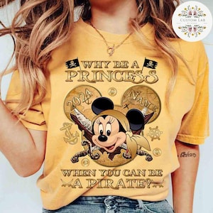 K&ouml;nnte beinhalten: Gelbes T-Shirt mit einer goldenen Grafik von Mickey Mouse, der als Pirat verkleidet ist. Die Grafik enth&auml;lt eine Schatzkarte, Schwerter und den Text "Warum eine Prinzessin sein, wenn man ein Pirat sein kann?"
