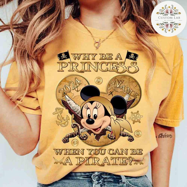 K&ouml;nnte beinhalten: Gelbes T-Shirt mit einer goldenen Grafik von Mickey Mouse, der als Pirat verkleidet ist. Die Grafik enth&auml;lt eine Schatzkarte, Schwerter und den Text "Warum eine Prinzessin sein, wenn man ein Pirat sein kann?"