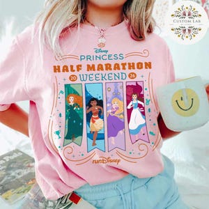 May include: A light pink t-shirt featuring the text "Disney Princess Half Marathon Weekend 26" with illustrations of Disney princesses. A white mug with a smiley face is held in the hand.