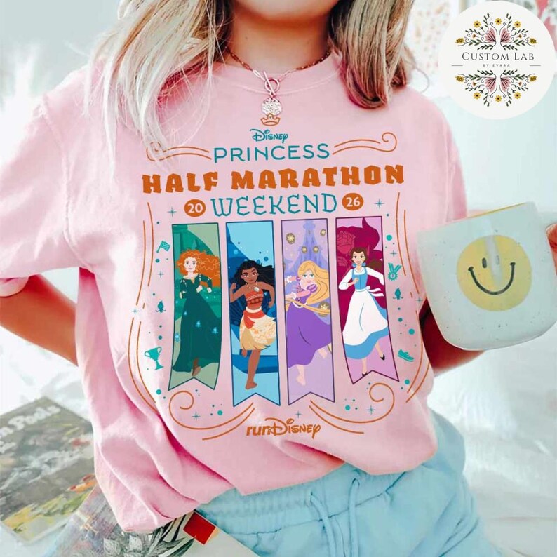 May include: A light pink t-shirt featuring the text "Disney Princess Half Marathon Weekend 26" with illustrations of Disney princesses. A white mug with a smiley face is held in the hand.