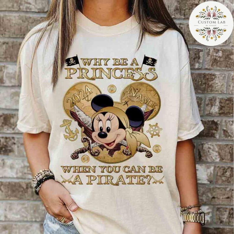 K&ouml;nnte beinhalten: Ein wei&szlig;es T-Shirt mit einer goldenen Grafik von Mickey Mouse, der als Pirat verkleidet ist. Die Grafik enth&auml;lt eine Schatzkarte, Schwerter und den Text "WHY BE A PRINCESS WHEN YOU CAN BE A PIRATE?"