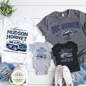 May include: A collection of t-shirts and a baby onesie featuring vintage car designs. The shirts are white, gray, and black, with the text "Fabulous Hudson Hornet," "Doc Hudson," and "Hudson Garage." The onesie is gray and has "Hudson Hornet" printed on it. The designs include a blue car.