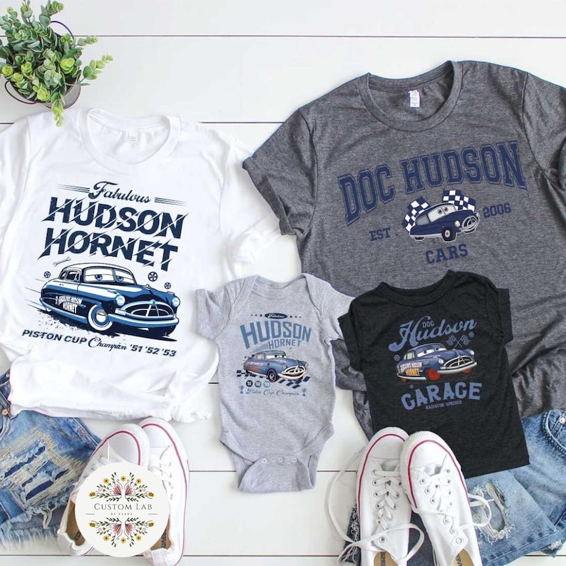 May include: A collection of t-shirts and a baby onesie featuring vintage car designs. The shirts are white, gray, and black, with the text "Fabulous Hudson Hornet," "Doc Hudson," and "Hudson Garage." The onesie is gray and has "Hudson Hornet" printed on it. The designs include a blue car.