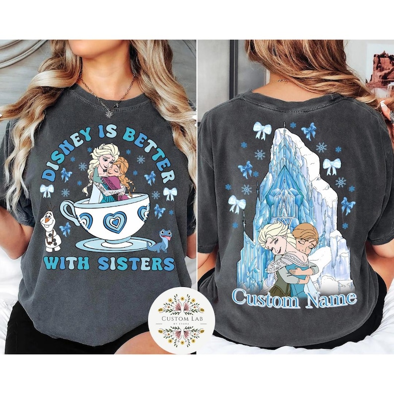 Puede incluir: Camiseta gris oscuro con el texto "DISNEY IS BETTER WITH SISTERS" y un gr&aacute;fico de Elsa y Anna en una taza de t&eacute;. La parte trasera muestra a las hermanas abraz&aacute;ndose frente a un castillo de hielo, con el texto "Custom Name".