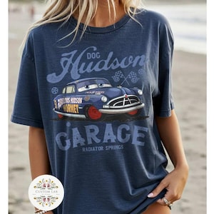 May include: Navy blue t-shirt featuring a graphic of a blue cartoon car with red wheels, the text "Hudson Garage" and "Radiator Springs". The shirt also has the text "Fabulous Hudson Hornet".