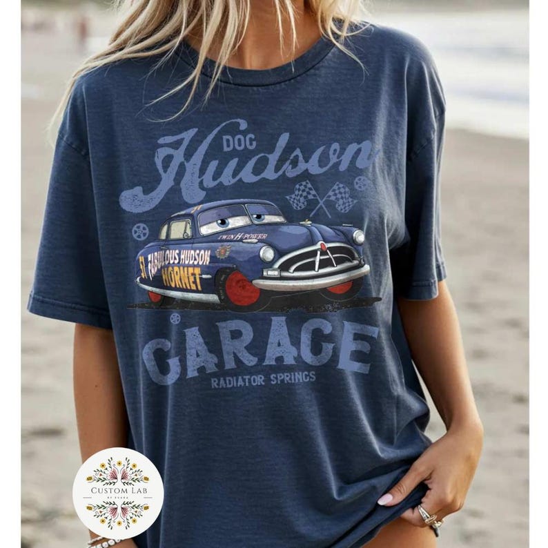 May include: Navy blue t-shirt featuring a graphic of a blue cartoon car with red wheels, the text "Hudson Garage" and "Radiator Springs". The shirt also has the text "Fabulous Hudson Hornet".
