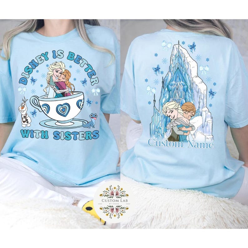 Puede incluir: Camiseta azul claro con tem&aacute;tica de Disney. La parte delantera presenta un gr&aacute;fico de Elsa y Anna en una taza de t&eacute; con el texto "Disney is Better With Sisters". La parte trasera muestra a las hermanas abraz&aacute;ndose frente a un castillo con el texto "Custom Name."