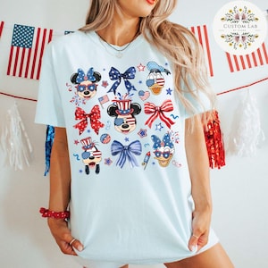 Peut inclure: Un t-shirt bleu clair avec un motif patriotique repr&eacute;sentant Mickey Mouse, Minnie Mouse, Donald Duck et Dingo portant des v&ecirc;tements et des accessoires rouges, blancs et bleus. Le motif comprend des &eacute;toiles, des n&oelig;uds et des feux d'artifice.
