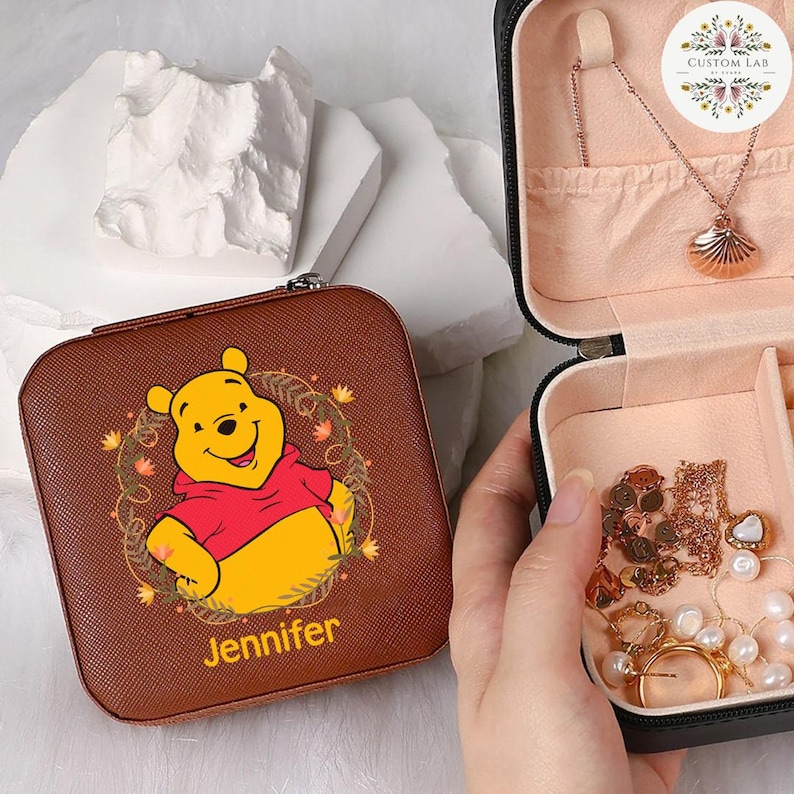 May include: A brown square jewelry case with a zipper closure. The case features a cartoon image of Winnie the Pooh wearing a red shirt, with the name "Jennifer" printed below. The case is open, revealing jewelry inside.