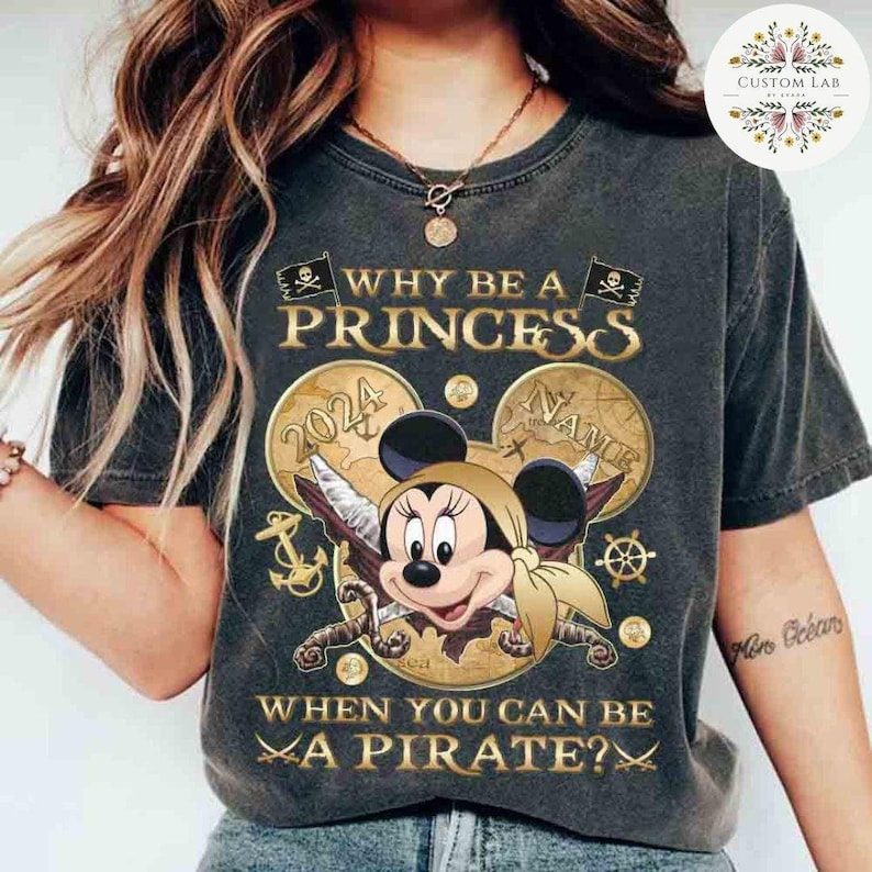 K&ouml;nnte beinhalten: Ein schwarzes T-Shirt mit Vintage-Look, das ein Cartoon-Bild von Minnie Mouse zeigt, die als Piratin verkleidet ist. Der Text "WHY BE A PRINCESS WHEN YOU CAN BE A PIRATE?" ist in Gold auf dem Shirt gedruckt. Das Bild enth&auml;lt eine Schatzkarte, einen Anker und einen Kompass.