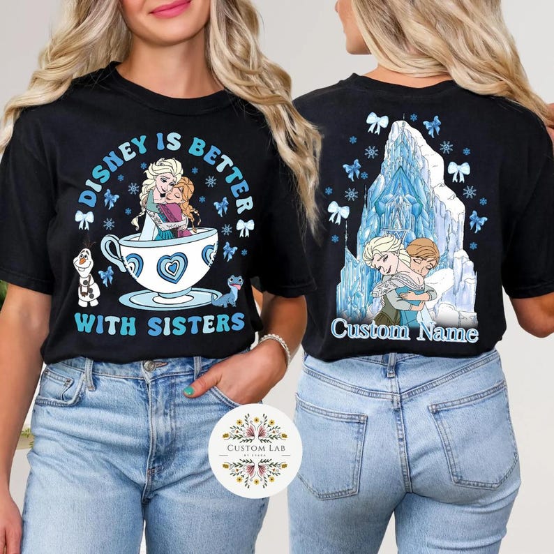 Puede incluir: Camiseta negra con el texto "DISNEY IS BETTER WITH SISTERS" en azul y blanco. El dise&ntilde;o frontal presenta a Elsa y Anna en una taza de t&eacute;. La parte trasera de la camiseta tiene un dise&ntilde;o de Elsa y Anna abraz&aacute;ndose frente a un castillo de hielo azul.
