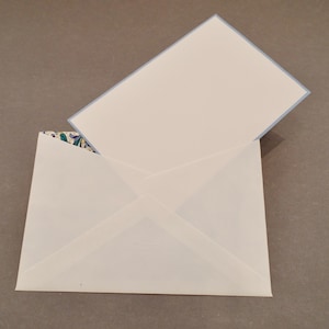 May include: A white envelope with a triangular flap and a patterned interior lining featuring blue, green, and yellow floral motifs. A matching white card with a light blue border is partially inserted into the envelope, which has the word Crane embossed.