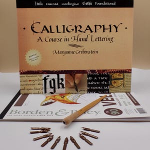 Pode incluir: Um conjunto de caligrafia com um livro intitulado "Calligraphy: A Course in Hand Lettering" de Maryanne Grebenstein. O conjunto inclui uma caneta de madeira, várias pontas e uma revista. O livro tem o texto "Italic, Uncial, Carolingian, Gothic, Foundational".