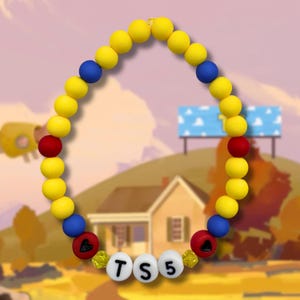 May include: A beaded bracelet featuring a repeating pattern of yellow, blue, and red spherical beads. The bracelet includes two red beads with black heart icons and three white cylindrical beads with the black letters T, S, and the number 5.