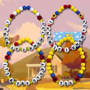 May include: Four circular beaded necklaces or bracelets featuring yellow, blue, red, and white beads. The white beads are printed with black letters spelling out the phrases I KNEW IT I KNEW IT and ITS A TAY STORY or TS5, interspersed with small yellow crystals.