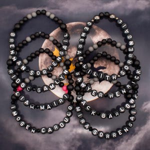 May include: A collection of black and grey beaded bracelets with white lettering spelling out names, including "Brennan", "Sorrengail", "Lilith", "Sawyer", "Jack", "Omri", "Cardulo", "Mair", "Liam", "Imogen", "Barlow", "Henrick", and "Gamlyn".