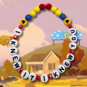 May include: A circular beaded necklace featuring white letter beads spelling out I KNEW IT I KNEW YOU. The design includes yellow, blue, and red spherical beads, small red faceted crystal beads, and two yellow beads with black heart symbols at the top.