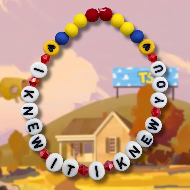 May include: A circular beaded necklace featuring white letter beads spelling out I KNEW IT I KNEW YOU. The design includes yellow, blue, and red spherical beads, small red faceted crystal beads, and two yellow beads with black heart symbols at the top.