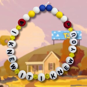 May include: A circular beaded bracelet featuring white spherical beads with black letters spelling out I KNEW IT I KNEW YOU. The design includes yellow, blue, and white round beads, two red beads with black heart symbols, and small yellow faceted beads.