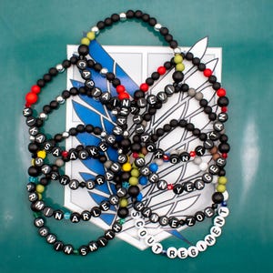 Op de afbeelding: Een set zwarte kralenarmbanden met witte letters die de namen van de personages uit de anime Attack on Titan spellen. De armbanden zijn in een cirkel gerangschikt op een groene achtergrond met een gedrukt beeld van het Attack on Titan-logo in het midden.