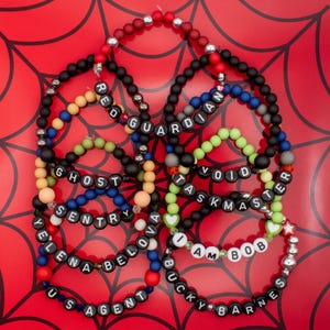 May include: A collection of beaded bracelets in various colors, including black, red, blue, and green. Each bracelet features white letter beads spelling out names like "RED GUARDIAN" and "TASKMASTER". The bracelets are arranged on a red background with a spiderweb design.