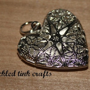 May include: Silver filigree heart charm pendant with a delicate openwork design. The heart is hollow and has a loop at the top for attaching to a chain or bracelet.