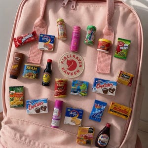 May include: A light pink backpack featuring a circular Fjällräven Kånken logo patch and numerous miniature replicas of various food and household product packages attached to the front, including Airheads, Spam, Hormel Chili, and Hostess CupCakes.
