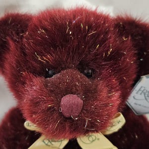 Puede incluir: Primer plano de un oso de peluche burdeos con una cinta amarilla clara atada en un lazo. El oso tiene una nariz burdeos oscuro y ojos negros. Purpurina dorada acentúa el pelaje. Una etiqueta con el nombre "Frango" está adjunta.