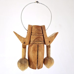 Puede incluir: Carrillón de viento de madera con acabado natural. El carillón presenta un cuerpo central con dos piezas laterales y dos piezas colgantes redondeadas. Se adjunta un lazo de alambre metálico para colgarlo. La madera tiene vetas visibles y un color marrón claro cálido.