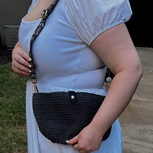 May include: A dark gray crocheted crossbody bag with a black strap featuring a floral pattern. The bag has a curved shape and a black leather snap closure. The strap is attached with gold-colored hardware.