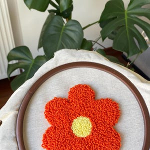 Op de afbeelding: Een close-up van een punch needle borduurproject met een fel oranje bloem met een gele kern. De borduursel is gespannen in een houten borduurring. De achtergrond bevat grote groene bladeren.