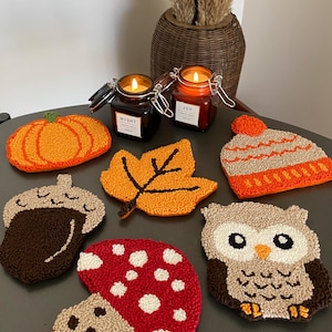 Könnte beinhalten: Ein Set aus sieben Herbst-Motiven, die mit der Punch-Needle-Technik hergestellt wurden. Die Motive umfassen einen Kürbis, eine Eichel, einen Pilz, ein Blatt, eine Eule und eine Mütze. Die Motive sind alle mit verschiedenen Garnfarben gefertigt und werden auf einem Holztisch präsentiert.
