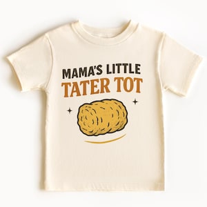 May include: Cream-colored t-shirt with the text "MAMA'S LITTLE TATER TOT" in brown and orange. A cartoon tater tot is below the text, with two small star graphics on either side. The shirt is a short-sleeved crew neck.