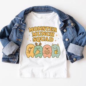 May include: White t-shirt with the text "MONSTER MUNCH SQUAD" in yellow and orange. The shirt features four cartoon monsters in different colours, each holding a snack. A blue denim jacket is draped over the shirt.