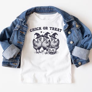 Peut inclure: T-shirt blanc avec un motif d'Halloween. Le motif représente trois poulets portant des chapeaux de sorcière et une citrouille d'Halloween, avec le texte "Chick or Treat" au-dessus. Une veste en jean bleue est drapée sur le t-shirt.